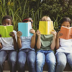 Image of a group of children sitting down and holding books in front of their faces. 