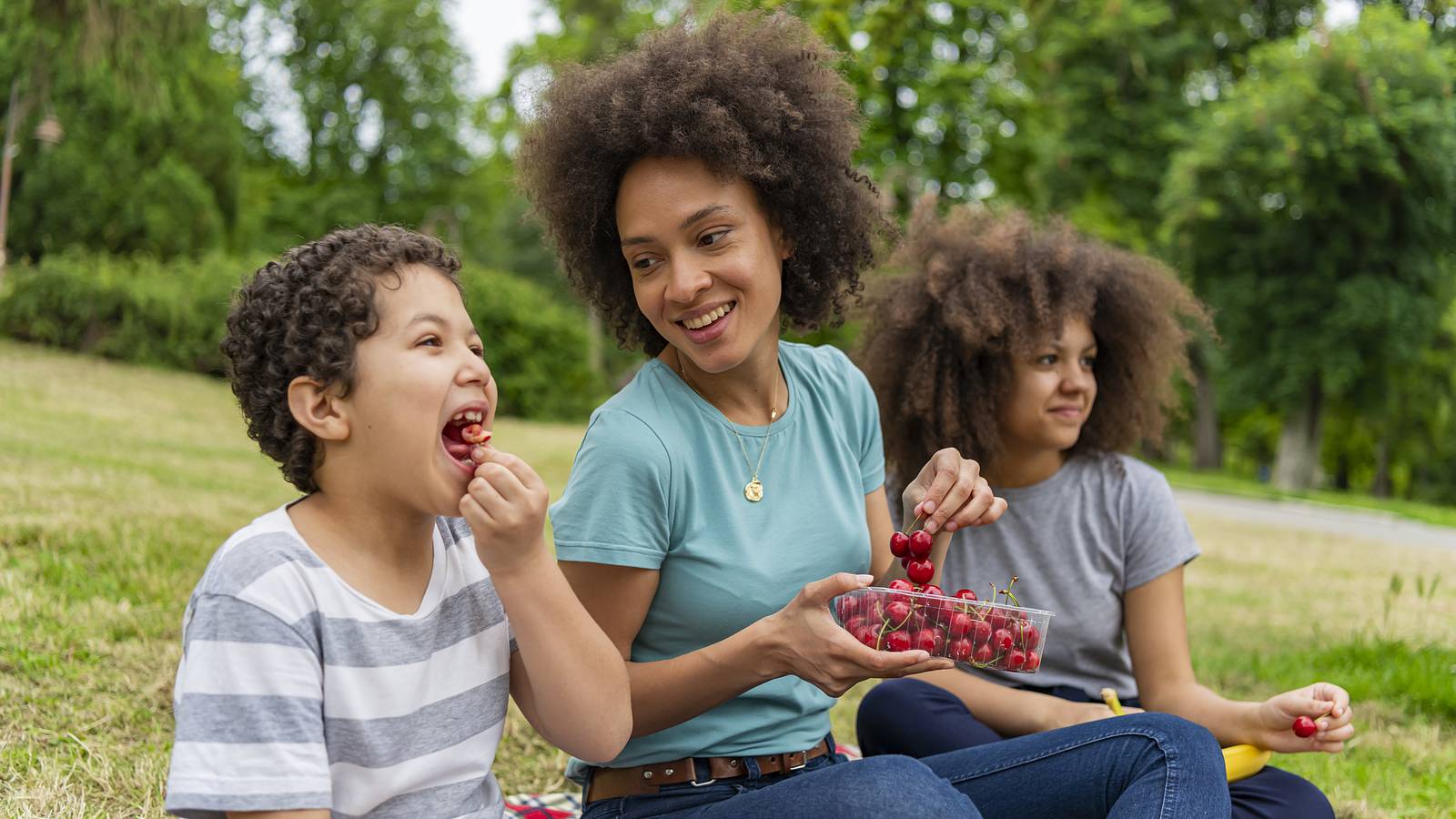 A mother and her two kids laugh while picnicking on cherries outside