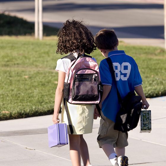 Image of a young girl and boy walking into school holding hands and lunchboxes. 